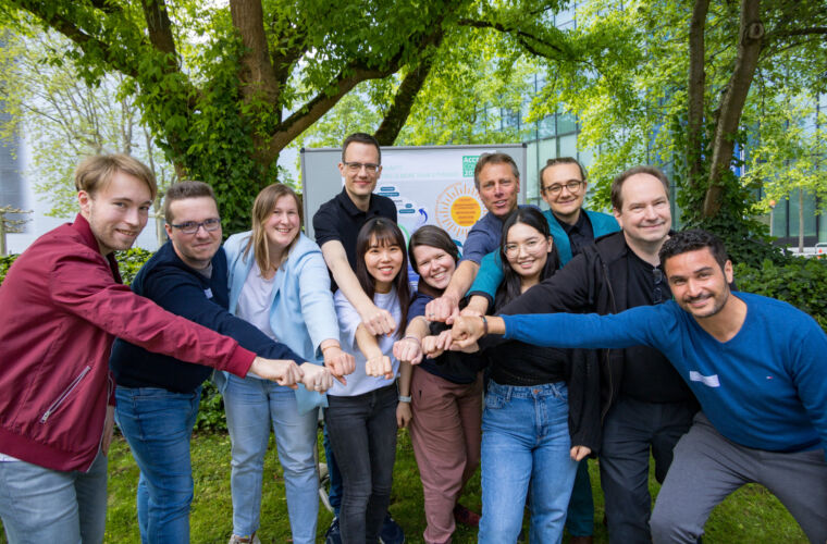 Die Test-Community steht im Freien unter Bäumen und streckt die Fäuste zusammen in die Mitte, als Zeichen von Zusammenhalt. Im Hintergrund ist ein Poster mit Diagrammen und Text zu sehen.