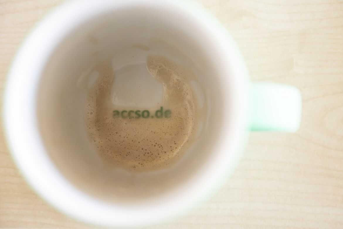 Ein Blick von oben in eine fast leere Kaffeetasse, die noch Reste von Schaum zeigt. Am Boden der Tasse ist der Schriftzug "accso.de" in grüner Schrift zu sehen. Die Tasse steht auf einer hellen Holzoberfläche.