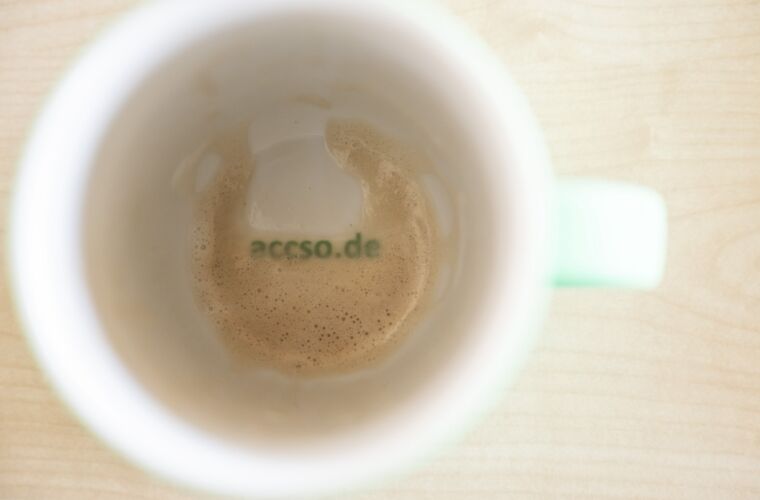 Ein Blick von oben in eine fast leere Kaffeetasse, die noch Reste von Schaum zeigt. Am Boden der Tasse ist der Schriftzug "accso.de" in grüner Schrift zu sehen. Die Tasse steht auf einer hellen Holzoberfläche.