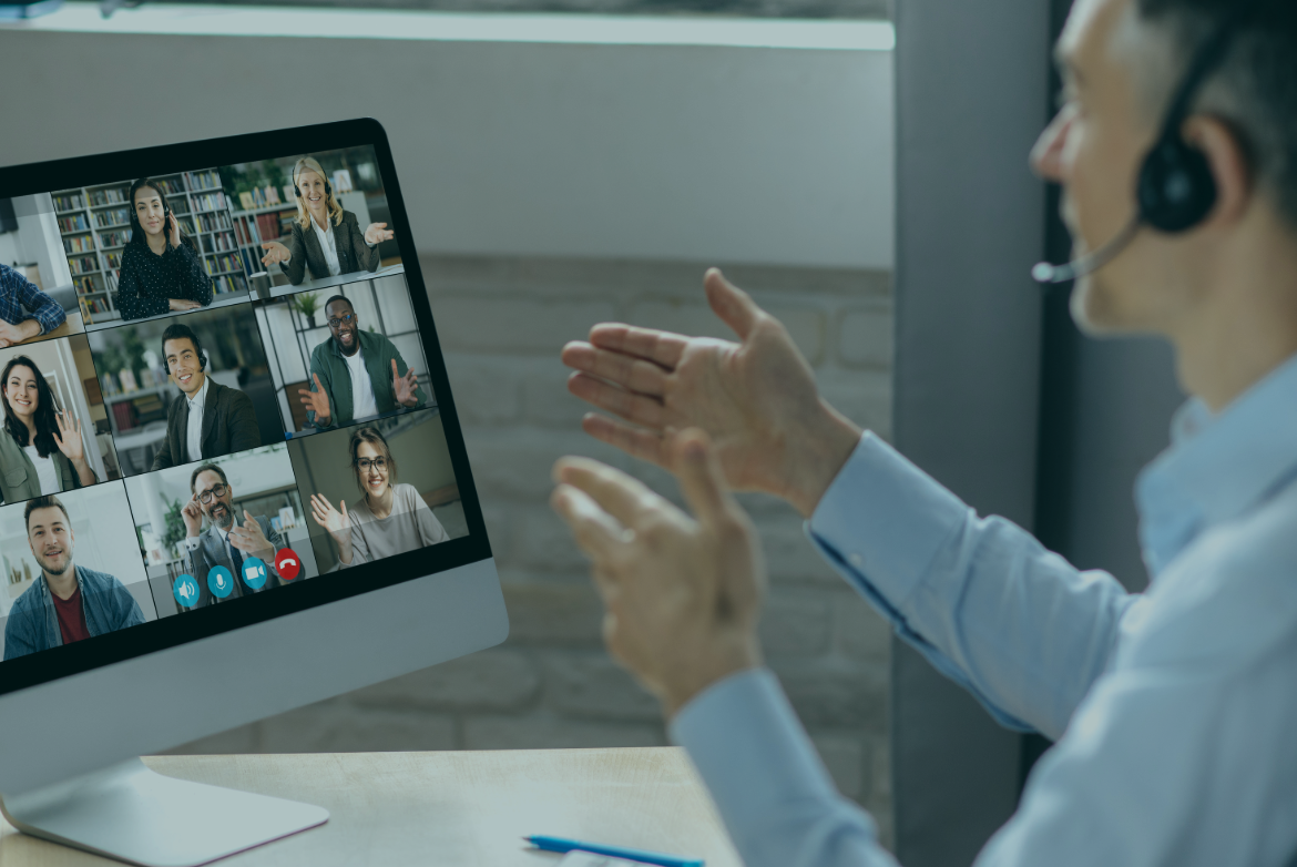 Person mit Headset spricht während einer Gruppen-Videokonferenz; Computerbildschirm zeigt mehrere Teilnehmende in einem Raster, einige winken oder gestikulieren.