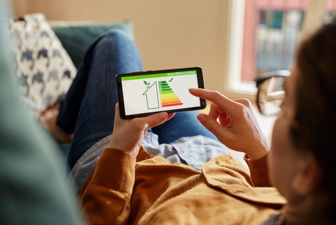 Eine Person liegt entspannt auf einem Sofa und hält ein Smartphone in der Hand. Auf dem Bildschirm des Smartphones ist eine Grafik mit einem Energieeffizienz-Label zu sehen, das die Energieeffizienz eines Hauses darstellt. Die Grafik zeigt eine farbige Skala von grün bis rot, die die Effizienzstufen anzeigt. Die Person berührt den Bildschirm, um die Informationen zu betrachten. Im Hintergrund ist ein gemütlicher Wohnraum mit einem Fenster zu erkennen.