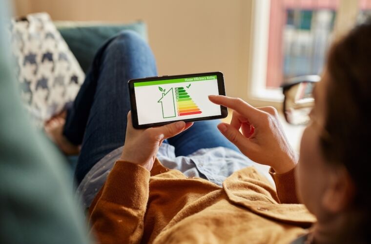 Eine Person liegt entspannt auf einem Sofa und hält ein Smartphone in der Hand. Auf dem Bildschirm des Smartphones ist eine Grafik mit einem Energieeffizienz-Label zu sehen, das die Energieeffizienz eines Hauses darstellt. Die Grafik zeigt eine farbige Skala von grün bis rot, die die Effizienzstufen anzeigt. Die Person berührt den Bildschirm, um die Informationen zu betrachten. Im Hintergrund ist ein gemütlicher Wohnraum mit einem Fenster zu erkennen.