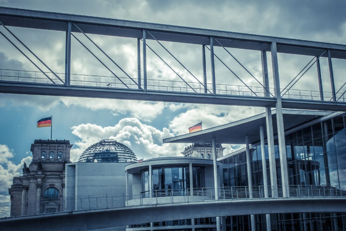 Moderne Glas- und Stahlarchitektur vor dem Reichstagsgebäude in Berlin, mit Glaskuppel, Brücke und Flaggen gegen einen wolkigen Himmel.