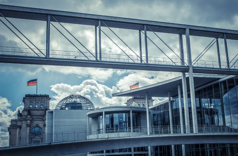 Moderne Glas- und Stahlarchitektur vor dem Reichstagsgebäude in Berlin, mit Glaskuppel, Brücke und Flaggen gegen einen wolkigen Himmel.