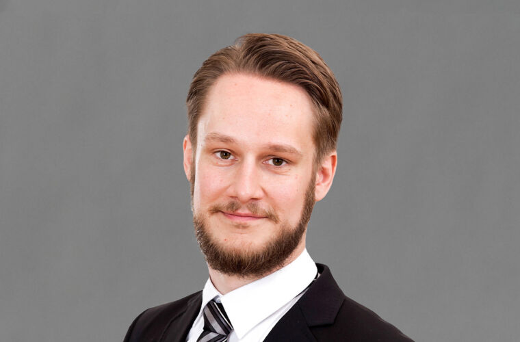 Junger Mann mit Vollbart und leicht geneigtem Kopf in die Kamera blickend. Er trägt einen Anzug, weißes Hemd und Krawatte.