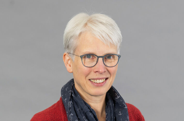 Porträt einer Frau mit weißen Haaren, Brille und dunklem Schal mit Anker-Muster. Sie trägt eine rote Strickjacke und lächelt.