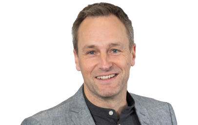 Eine Person mit kurzen braunen Haaren, grauem Blazer und dunklem Oberteil, lächelnd vor weißem Hintergrund, frontal aufgenommen.