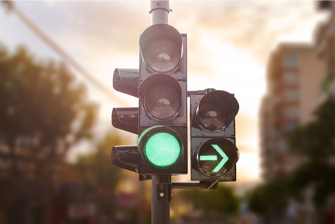 Eine Verkehrsampel zeigt ein grünes Licht und einen grünen Pfeil, der nach rechts weist, was darauf hinweist, dass der Verkehr in diese Richtung fließen darf. Der Hintergrund ist unscharf, mit angedeuteten Gebäuden und Bäumen, was auf eine städtische Umgebung hinweist.