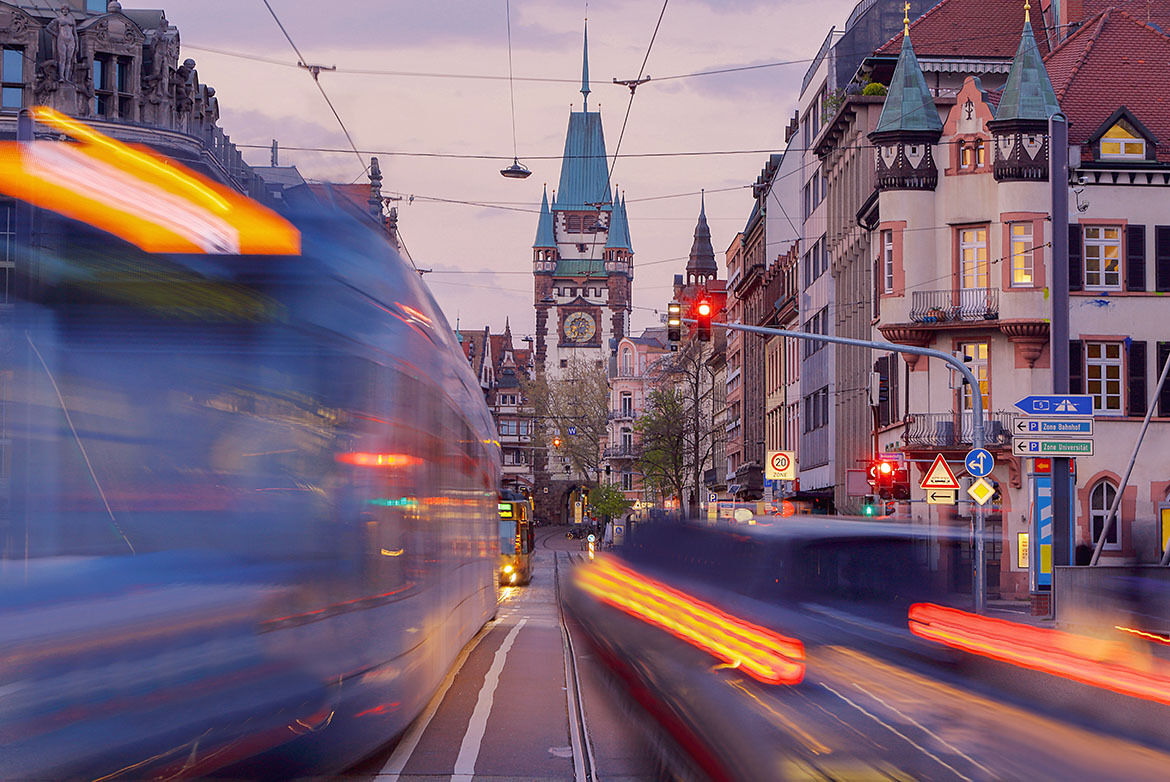Eine belebte Straßenszene in der Dämmerung zeigt eine Straßenbahn und ein Auto in Bewegung, deren Lichter in langen Streifen verwischen. Im Hintergrund ist ein historisches Tor mit einem Uhrturm zu sehen, das von alten Gebäuden mit verzierten Fassaden flankiert wird. Die Ampeln leuchten rot, und Straßenschilder weisen auf eine Zone mit Geschwindigkeitsbegrenzung hin.