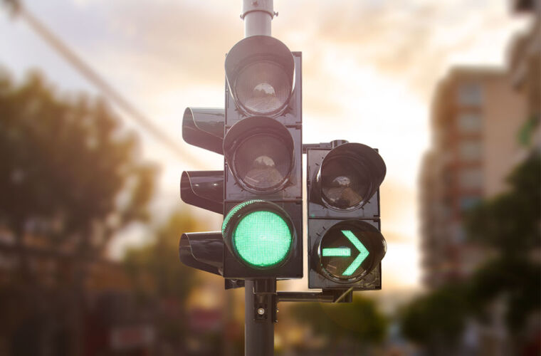 Eine Verkehrsampel zeigt ein grünes Licht und einen grünen Pfeil, der nach rechts weist, was darauf hinweist, dass der Verkehr in diese Richtung fließen darf. Der Hintergrund ist unscharf, mit angedeuteten Gebäuden und Bäumen, was auf eine städtische Umgebung hinweist.