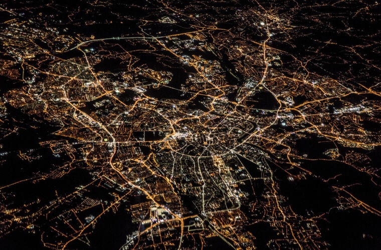 Eine nächtliche Luftaufnahme einer Stadt, die von einem Netz aus Straßen und beleuchteten Gebäuden durchzogen ist. Die Lichter der Stadt bilden ein komplexes Muster aus gelben und weißen Linien, die die Straßen und Gebäudeumrisse nachzeichnen. Dunkle Bereiche zwischen den beleuchteten Zonen zeigen unbebaute oder weniger beleuchtete Gebiete.