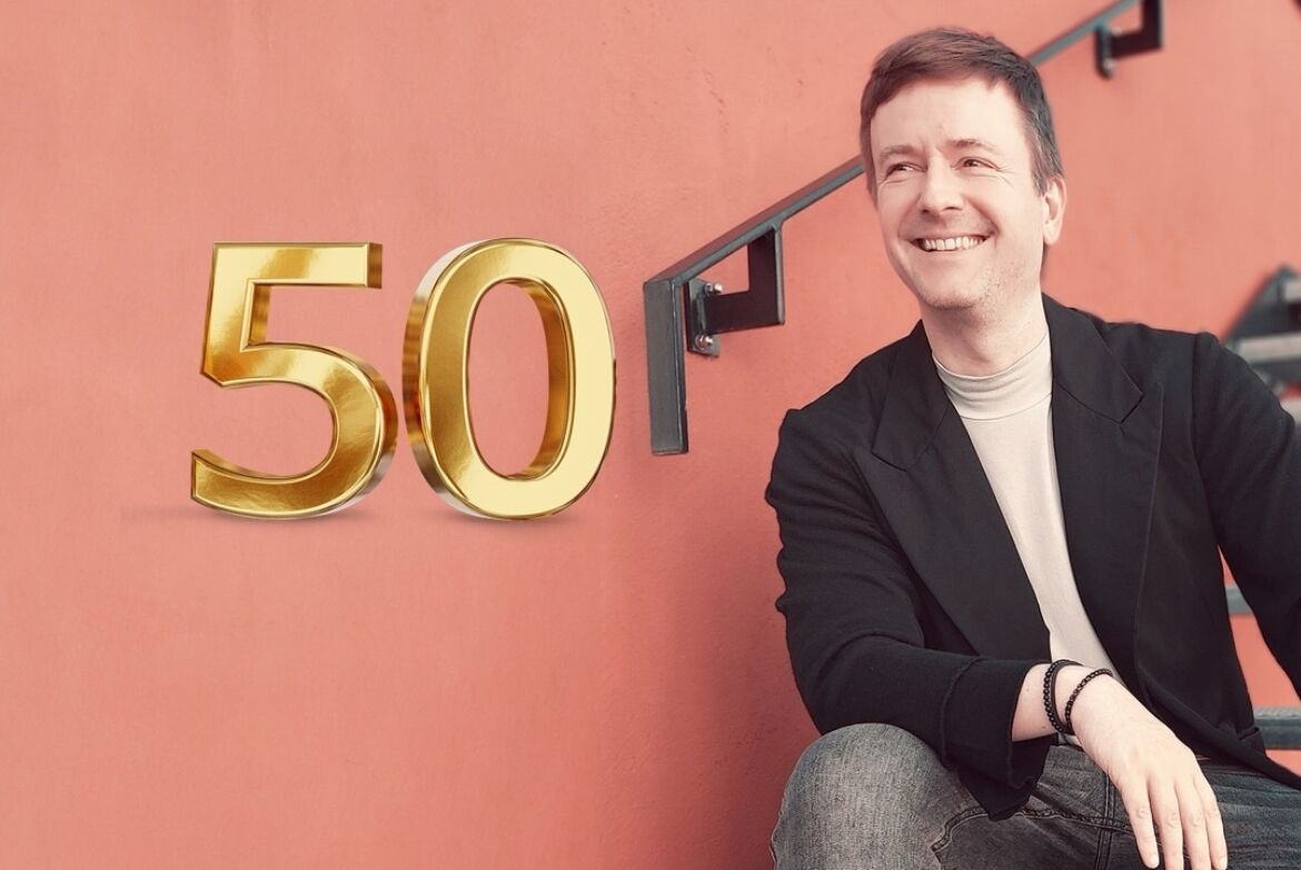 Marc Riedinger sitzt auf einer Treppe vor einer roten Wand, lächelnd. Links im Bild ist eine 50 zu sehen, die auf den 50. Artikel der Coaching@Accso Reihe hinweisen soll