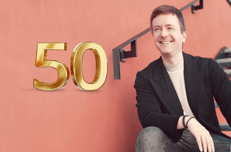 Marc Riedinger sitzt auf einer Treppe vor einer roten Wand, lächelnd. Links im Bild ist eine 50 zu sehen, die auf den 50. Artikel der Coaching@Accso Reihe hinweisen soll