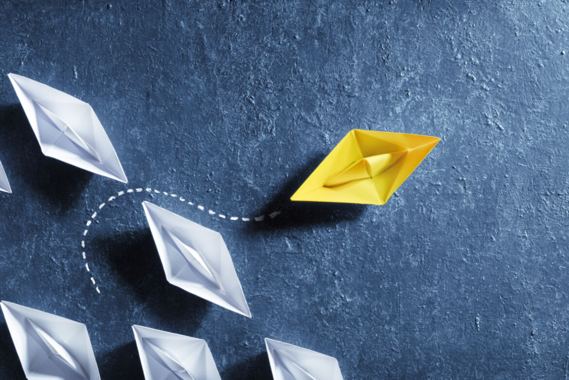Eine Gruppe aus weißen, gefalteten Papierbooten schwimmt in eine Richtung auf einer dunklen, strukturierten Oberfläche. Ein einzelnes gelbes Papierboot verlässt die Formation und bewegt sich in eine andere Richtung, angedeutet durch eine gestrichelte Linie. Das Bild symbolisiert Individualität, Innovation und das Brechen mit Konventionen.