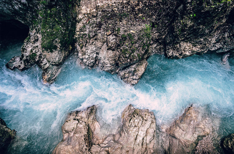 Ein türkisblauer Fluss fließt durch eine enge Schlucht aus grauen, felsigen Klippen. Das Wasser strömt schnell und schäumt weiß auf, während es sich seinen Weg zwischen den Felsen bahnt. Die umliegenden Felsen sind teilweise mit Moos und kleinen Pflanzen bewachsen.