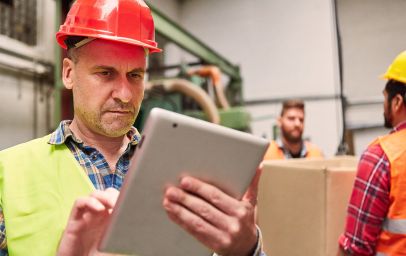 Eine Person in einem industriellen Umfeld trägt einen roten Schutzhelm und eine gelbe Warnweste. Sie hält ein Tablet in den Händen und scheint darauf zu arbeiten oder Informationen abzurufen. Im Hintergrund sind weitere Arbeiter in orangefarbenen Westen zu sehen, die mit Kartons beschäftigt sind.